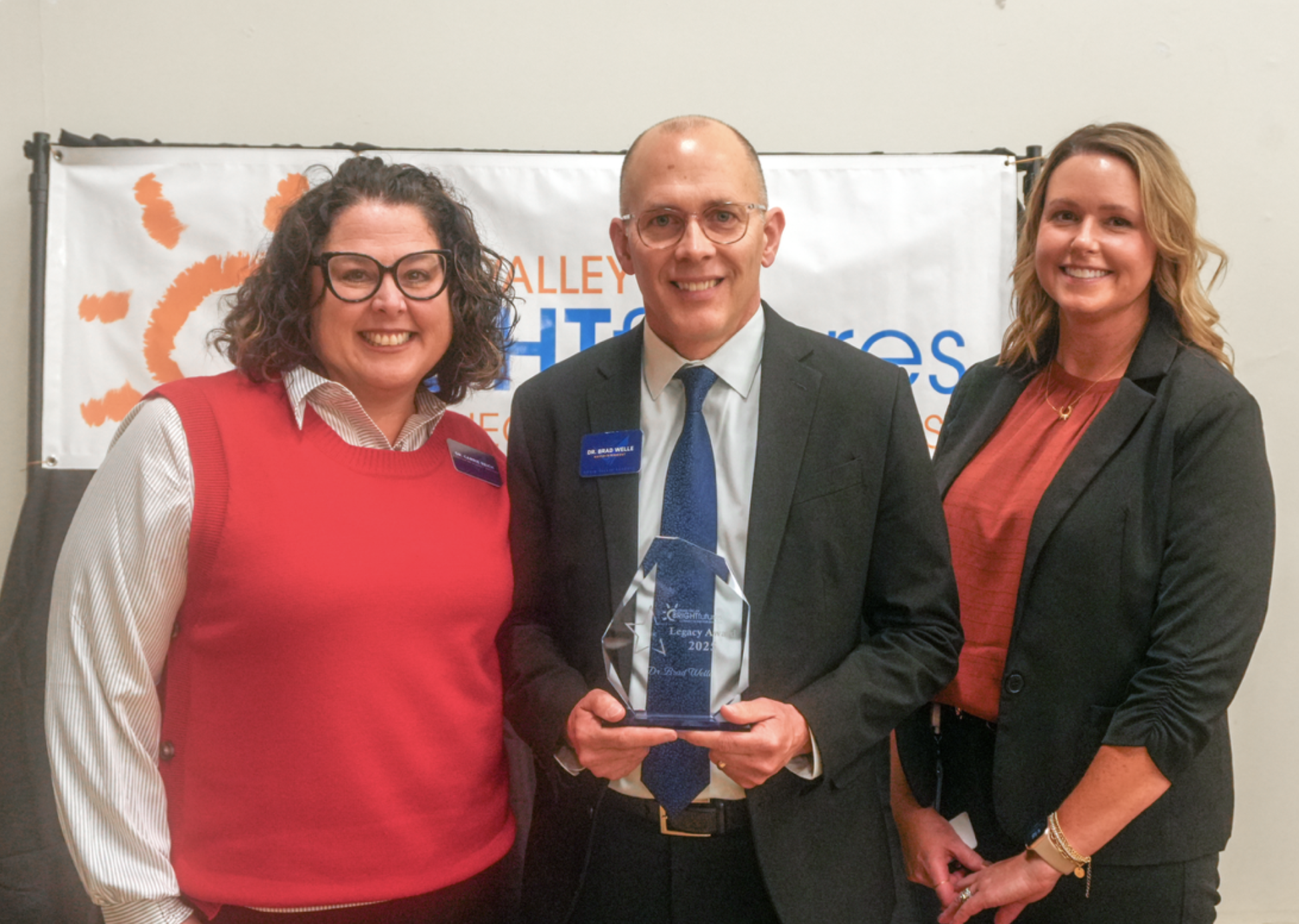 One individual holding an award with two other people standing on either side.They are in front of a banner that reads "Grain Valley Bright Futures". 