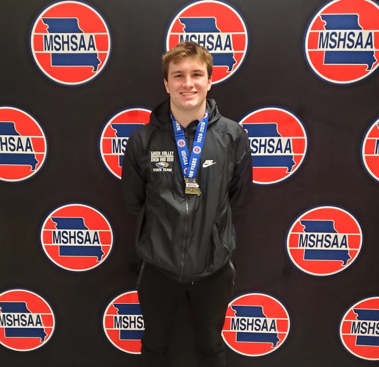 A photo of Gage Wright wearing a first place gold medal. Gage is standing in front of a photo background that has the MSHSAA logo repeating on a black background. 