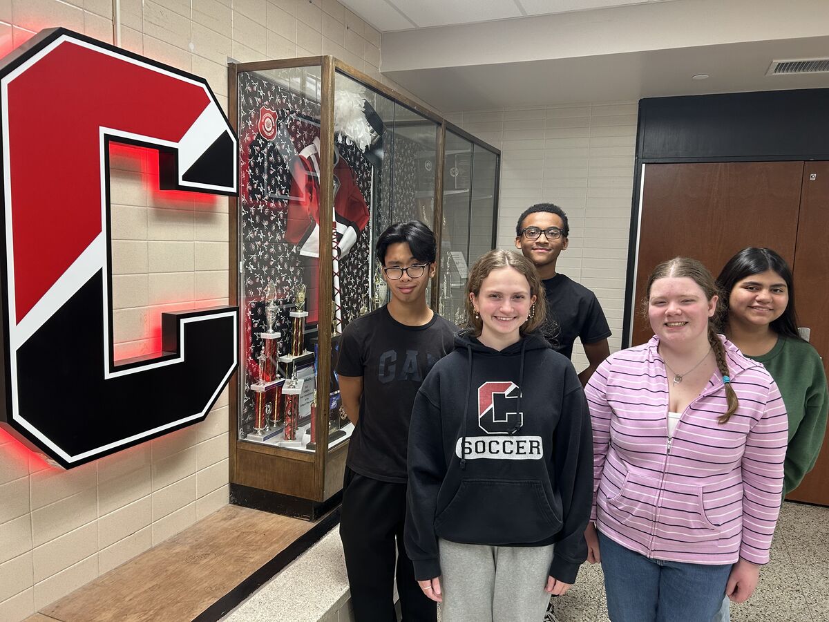Five students pose in a school hallway near a display case and a large letter 'C'.