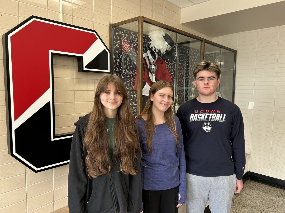 Three students pose in front of a school display case and a large letter 'C'.