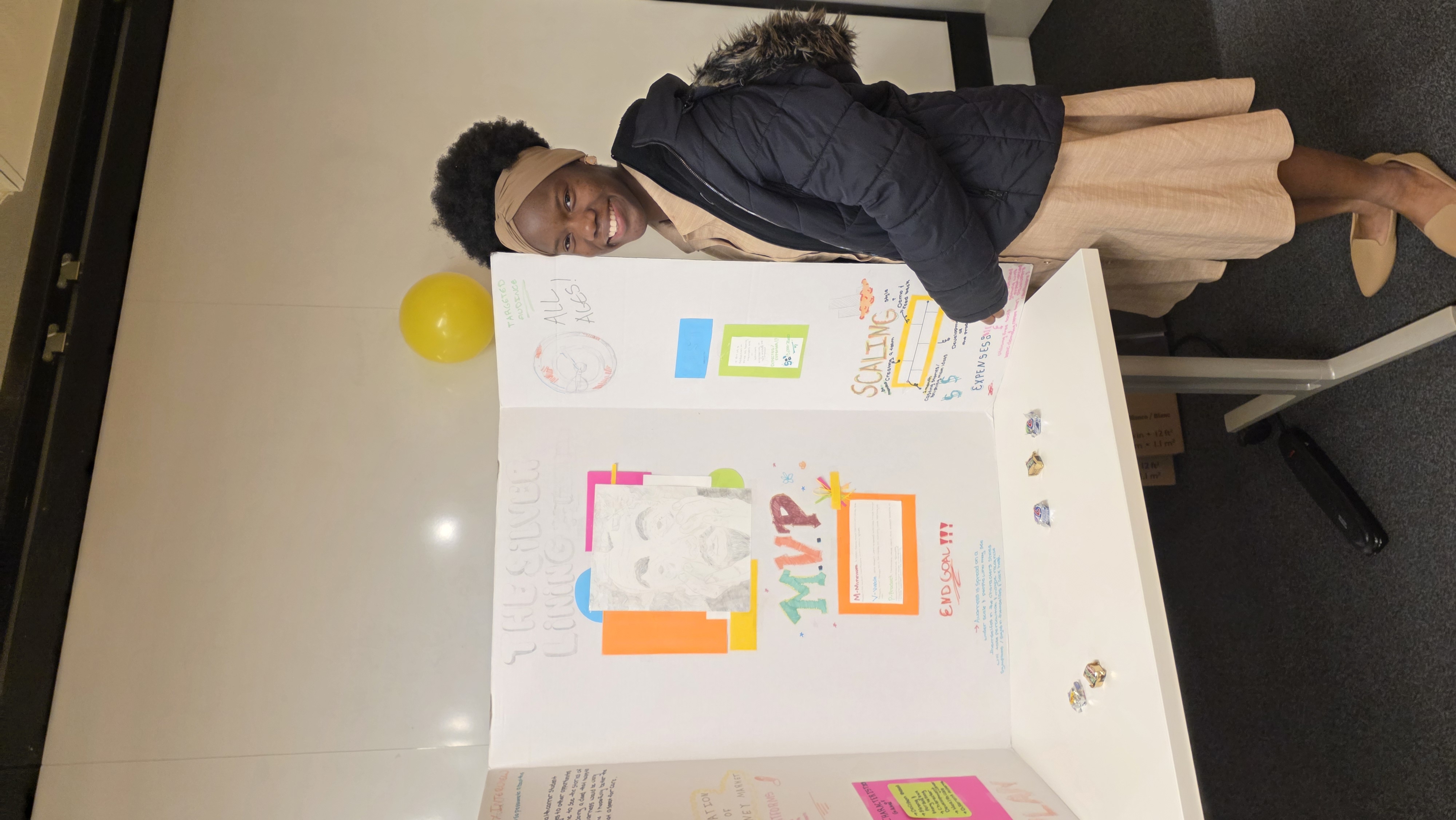 A young woman smiles, standing next to a presentation board with colorful notes and drawings.