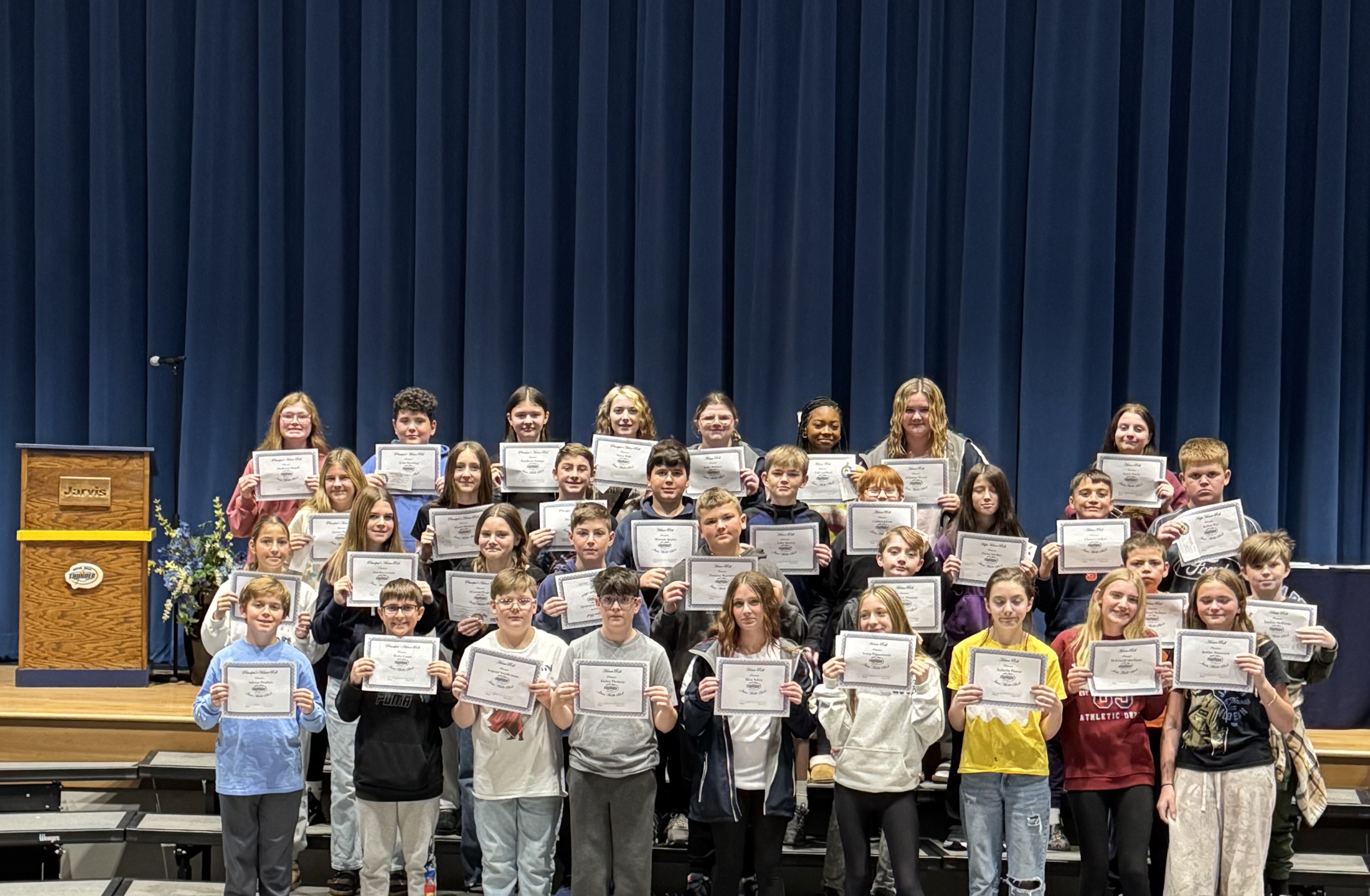 A group of students stand together, holding certificates in a school auditorium.