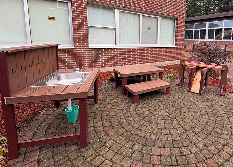 Outdoor play area with a sink, tables, and a water channel on a brick patio.