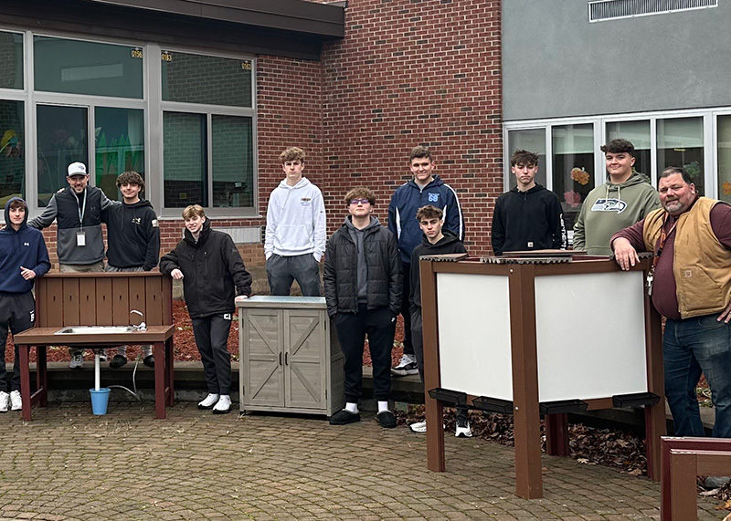 A group of students and instructors pose with outdoor projects in front of a brick building.