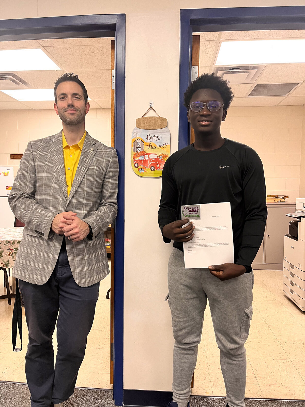 Two men stand in a doorway, one holding a document, smiling at the camera.
