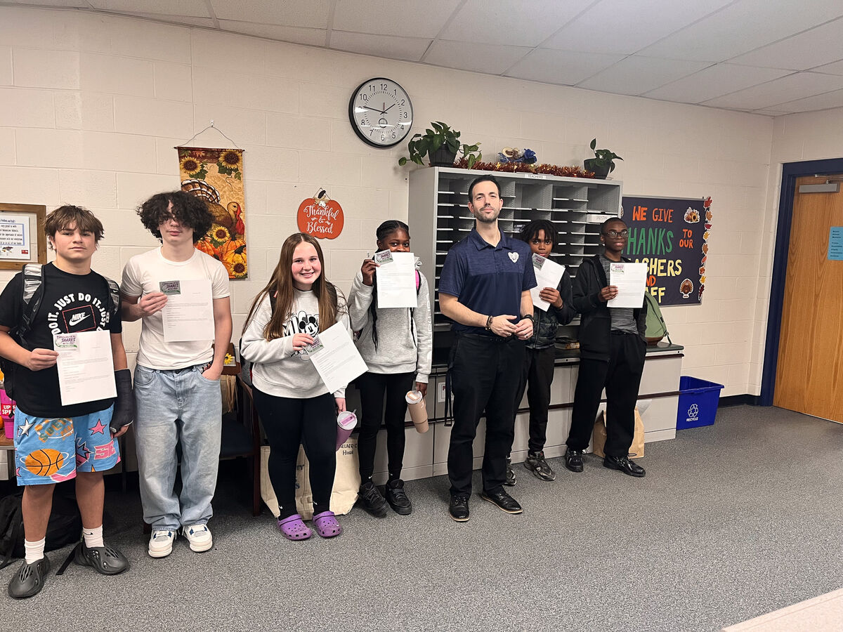 A group of students and an adult stand in a school hallway, holding papers.