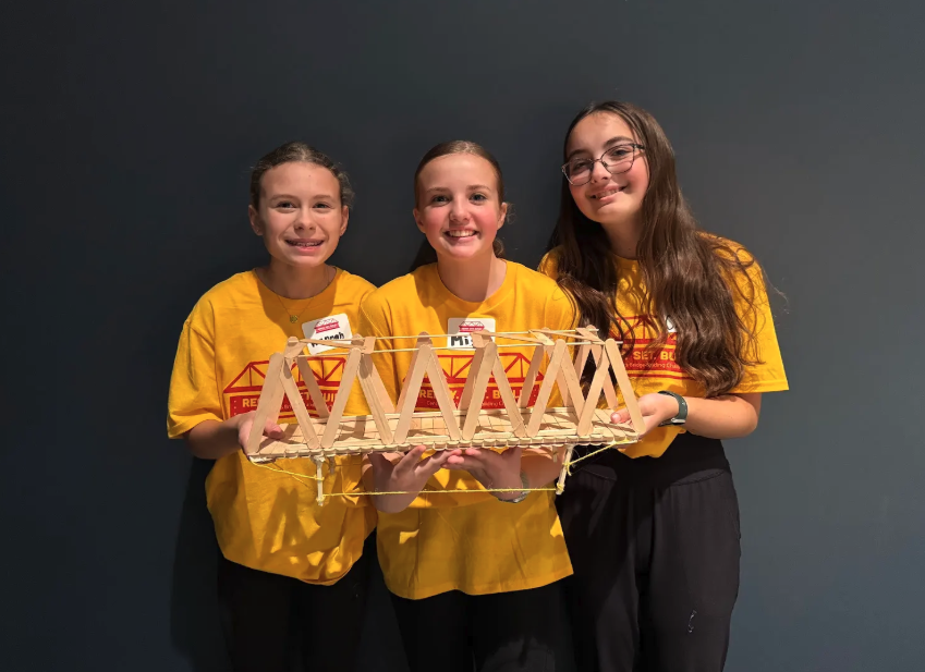 Three smiling girls hold up a popsicle stick bridge, posing for a photo.