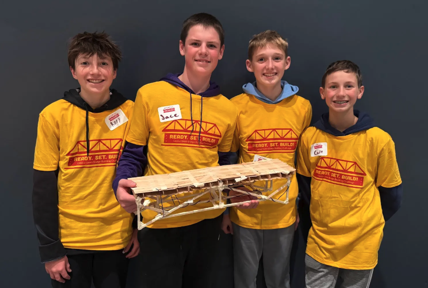 Four smiling boys pose with a model bridge, wearing matching yellow shirts.