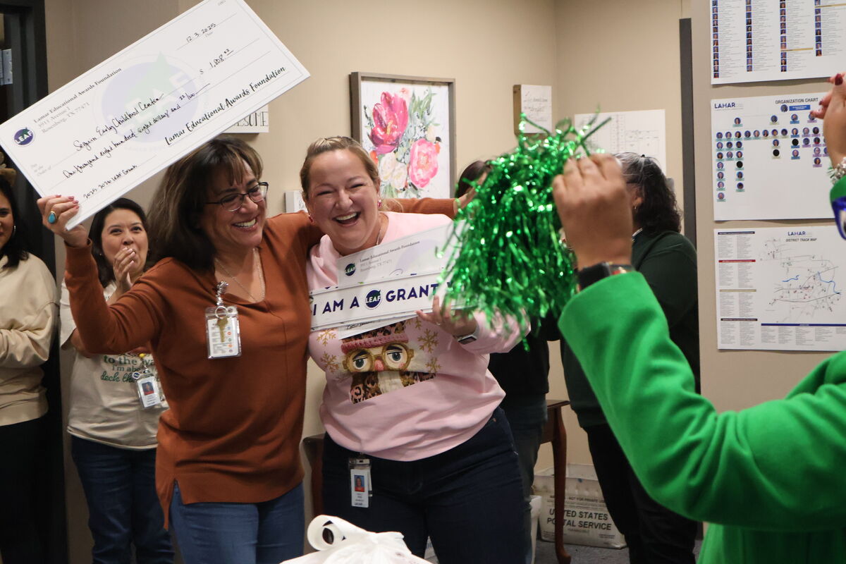 Two women celebrate with a large check and 'I am a Grant' signs, surrounded by others.