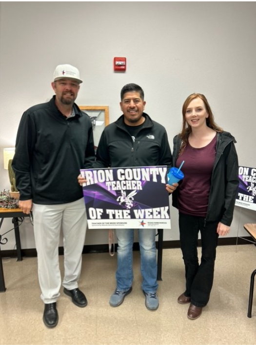 Three people pose for a photo, one holding a 'Teacher of the Week' sign.