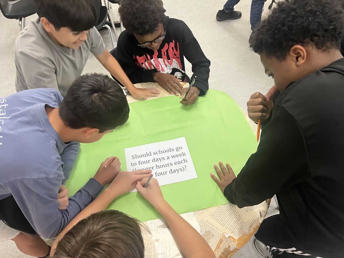 A group of students collaborate, writing on a green surface with a question about school schedules.