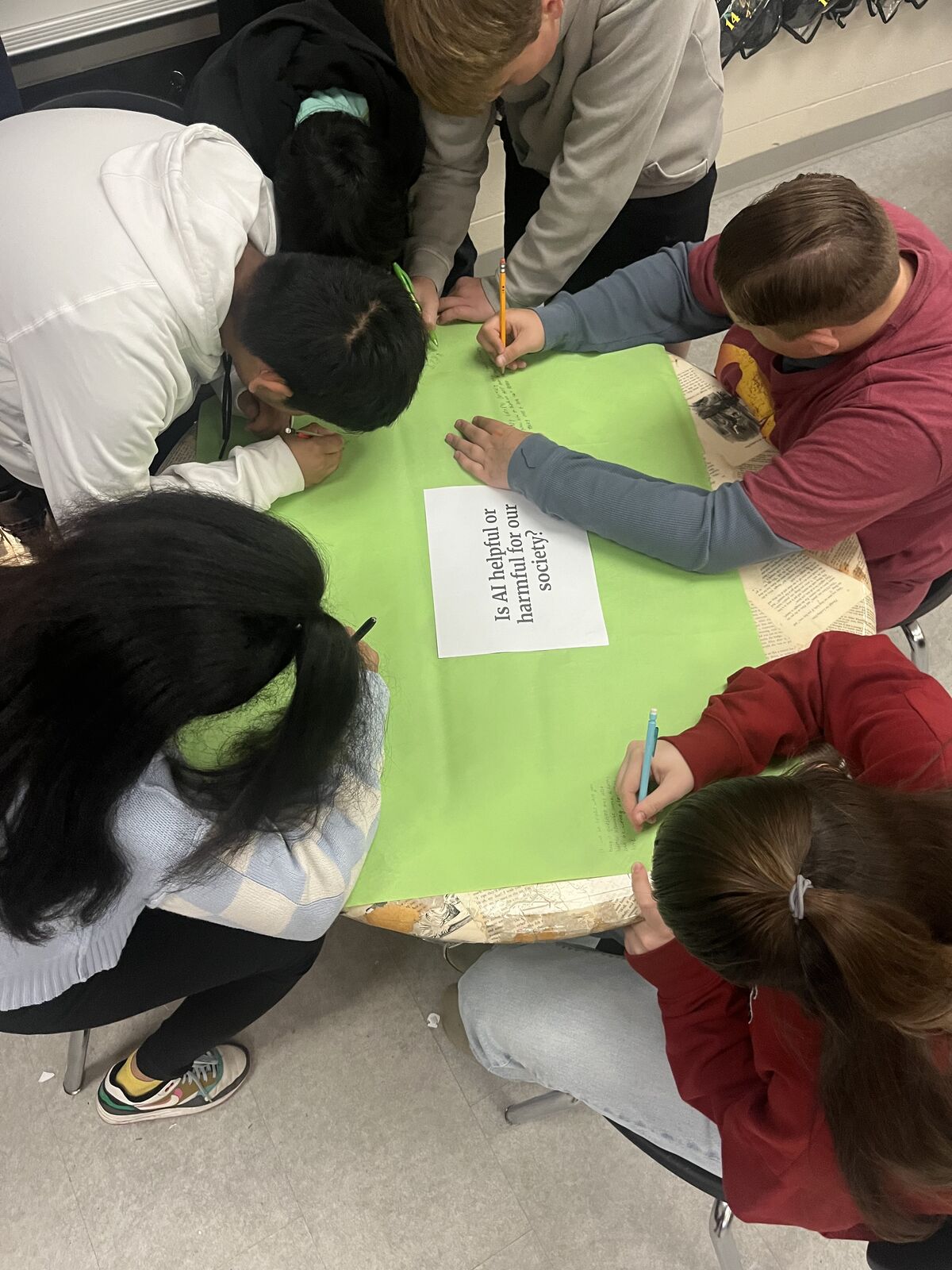 Students collaborate around a table, writing on a green poster with a question about AI.