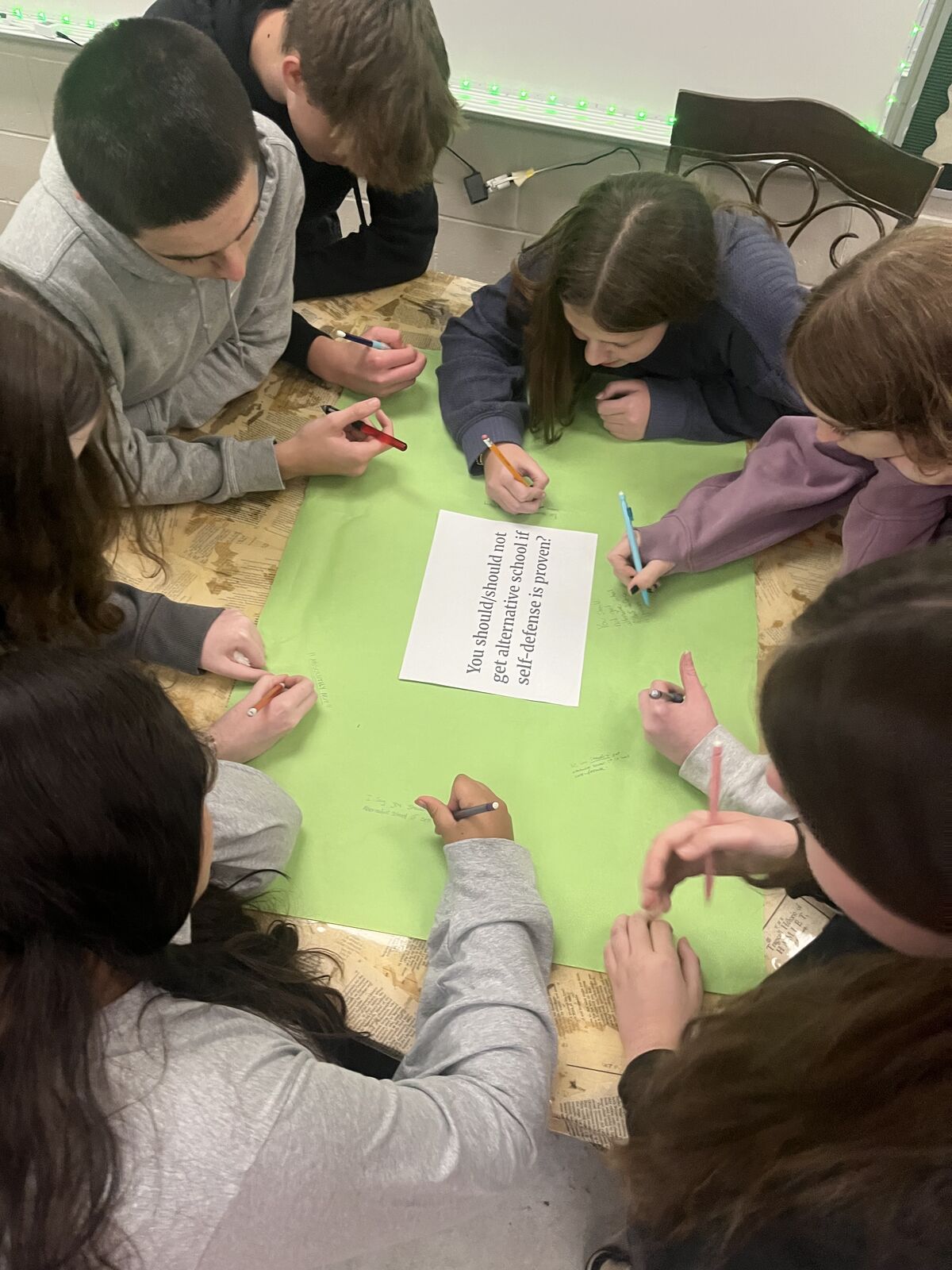 A group of students collaborate, writing on a green paper with a debate prompt.