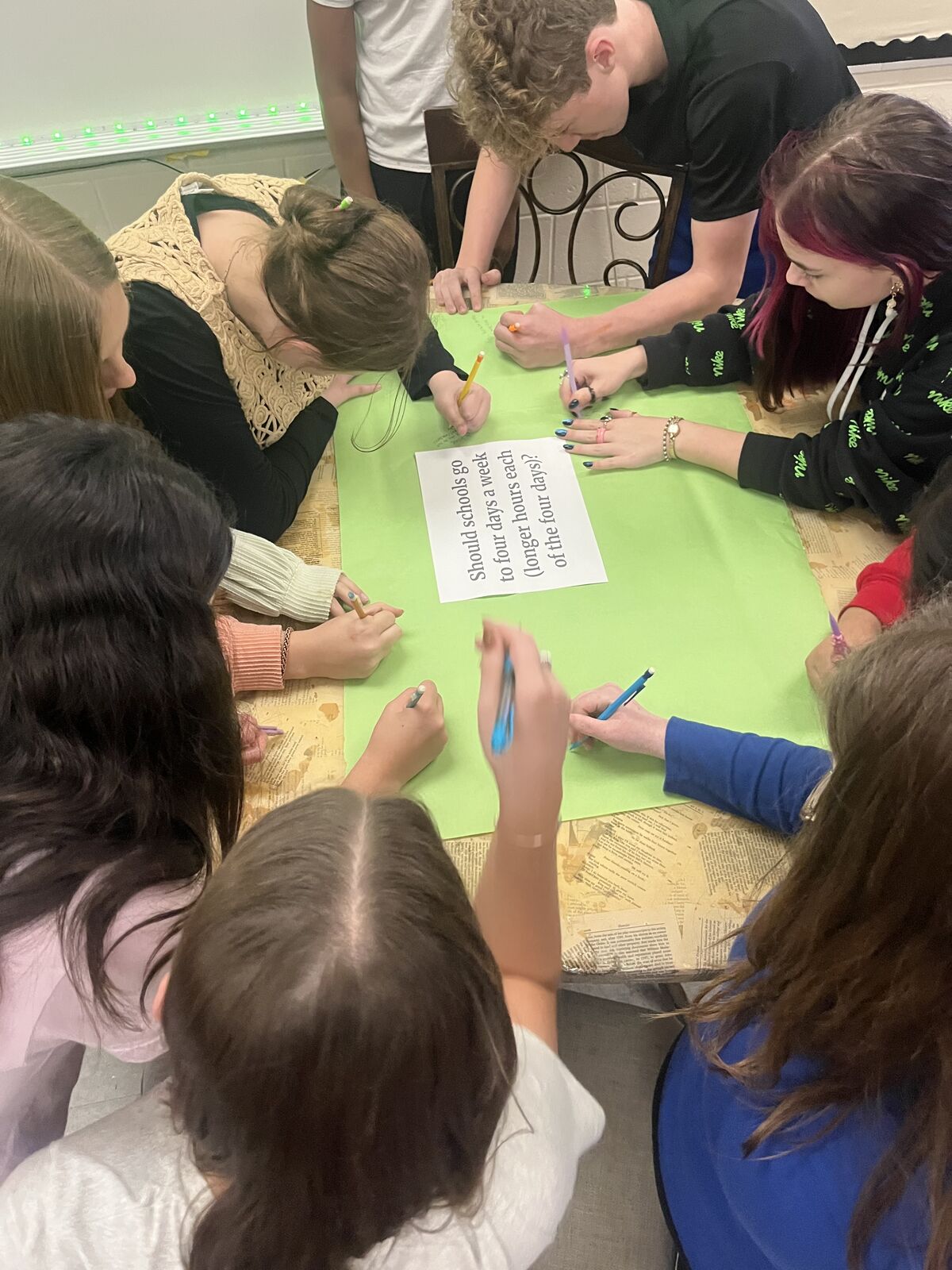 A group of people write on a green surface, discussing a school schedule change.