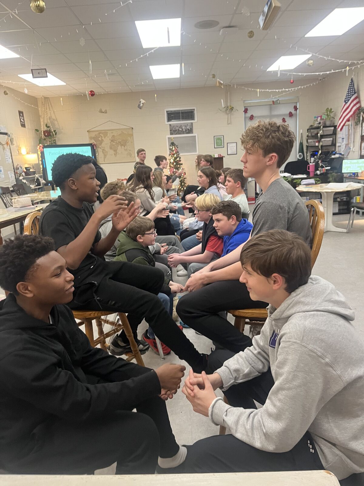 A group of students sit in a classroom, some with hands clasped together.
