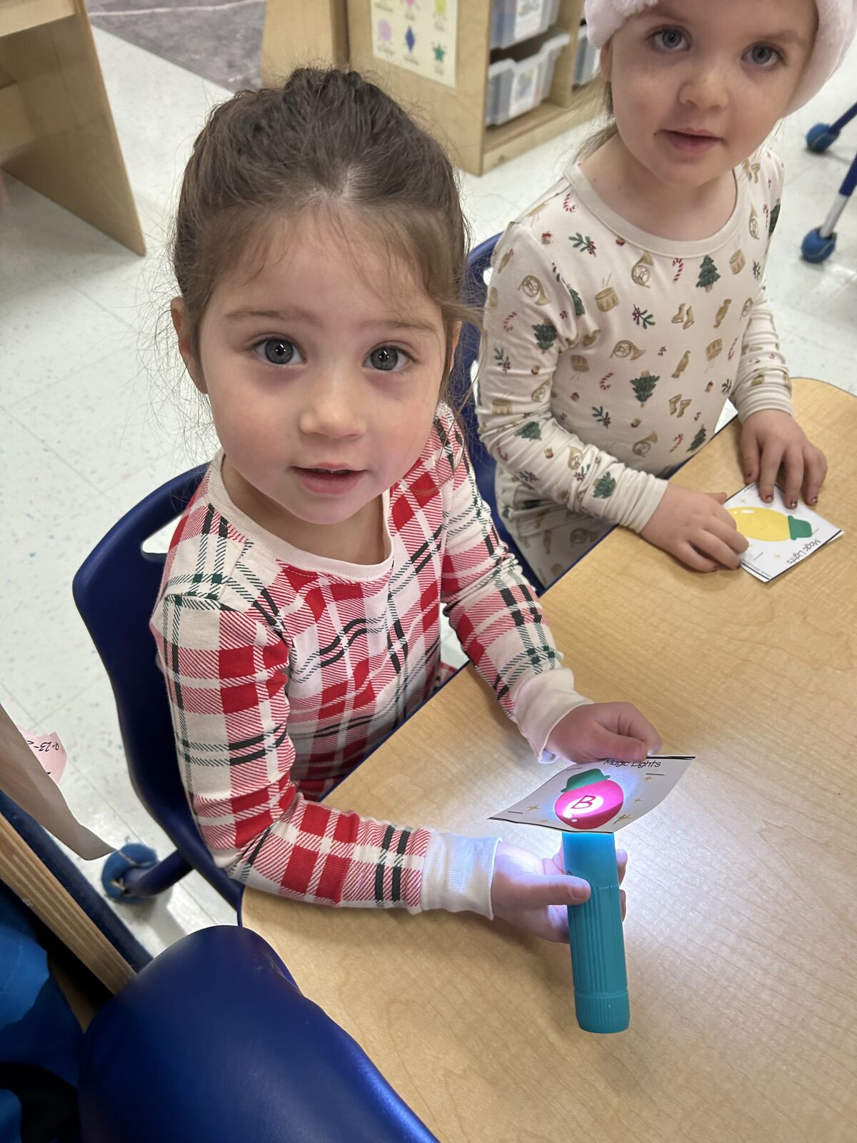 Two young children sit at a table, one holding a flashlight and a card.