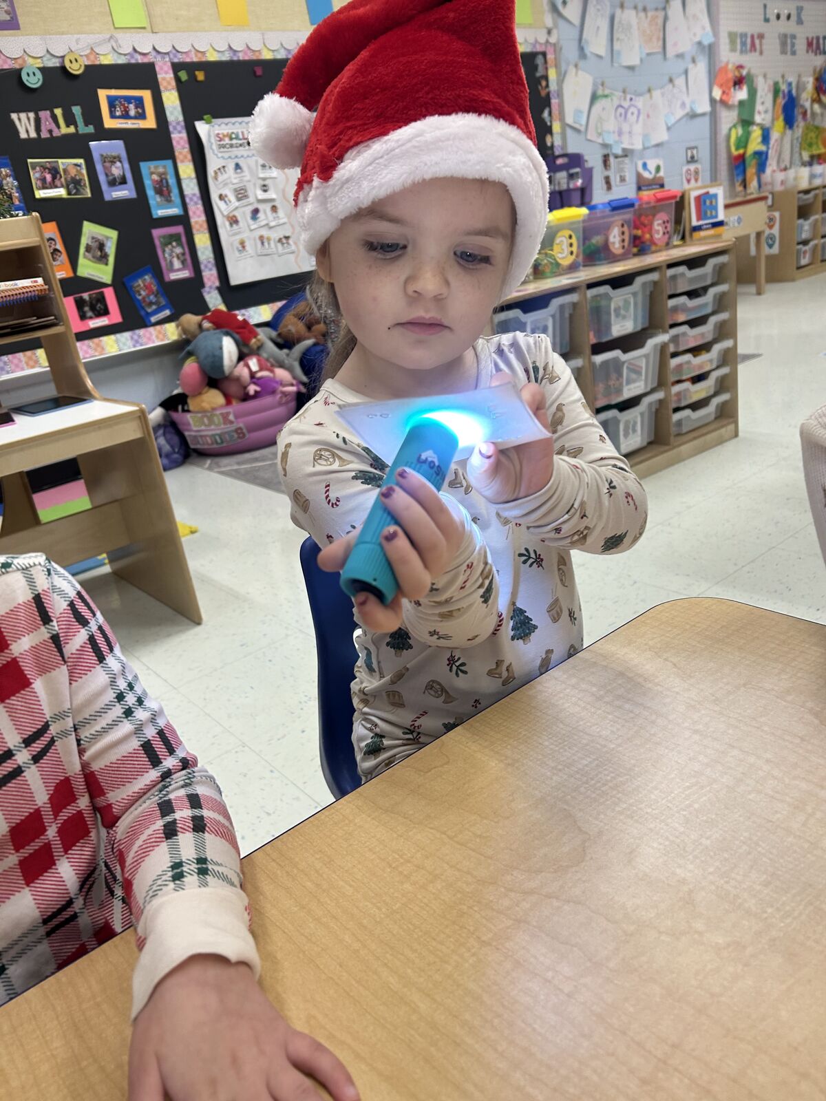 A young child wearing a Santa hat shines a flashlight at a table.