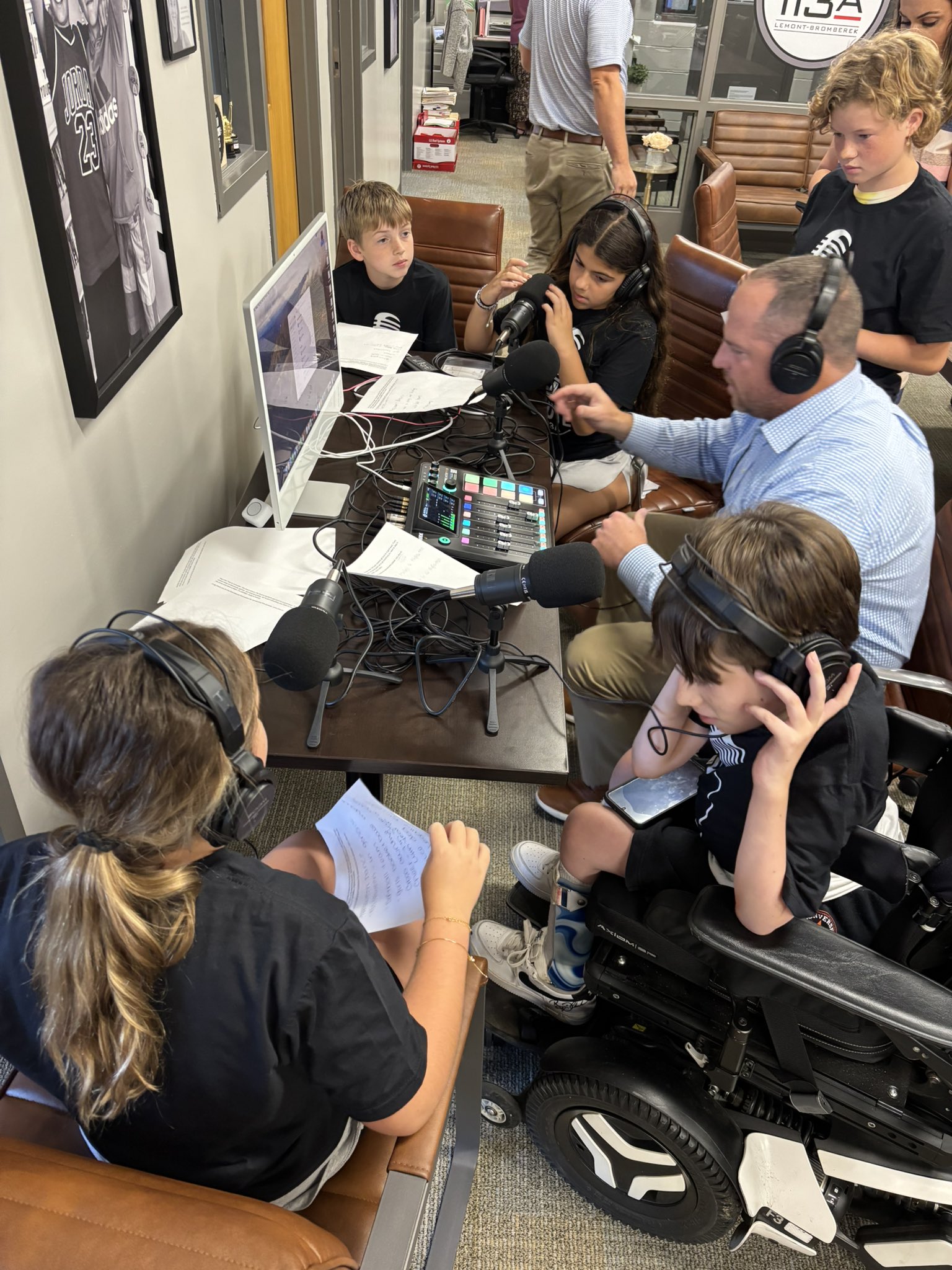 A group of children and an adult wearing headphones are gathered around a table with microphones and a soundboard, appearing to record a podcast.