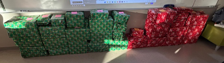A large collection of gifts wrapped in green and red patterned paper are stacked against a wall.
