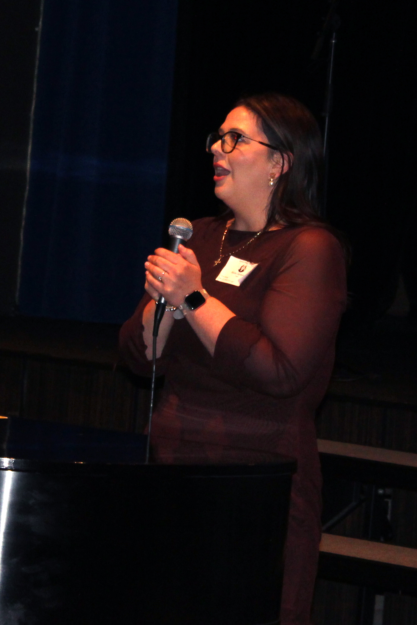A woman wearing glasses and a maroon top speaks into a microphone on a stage.