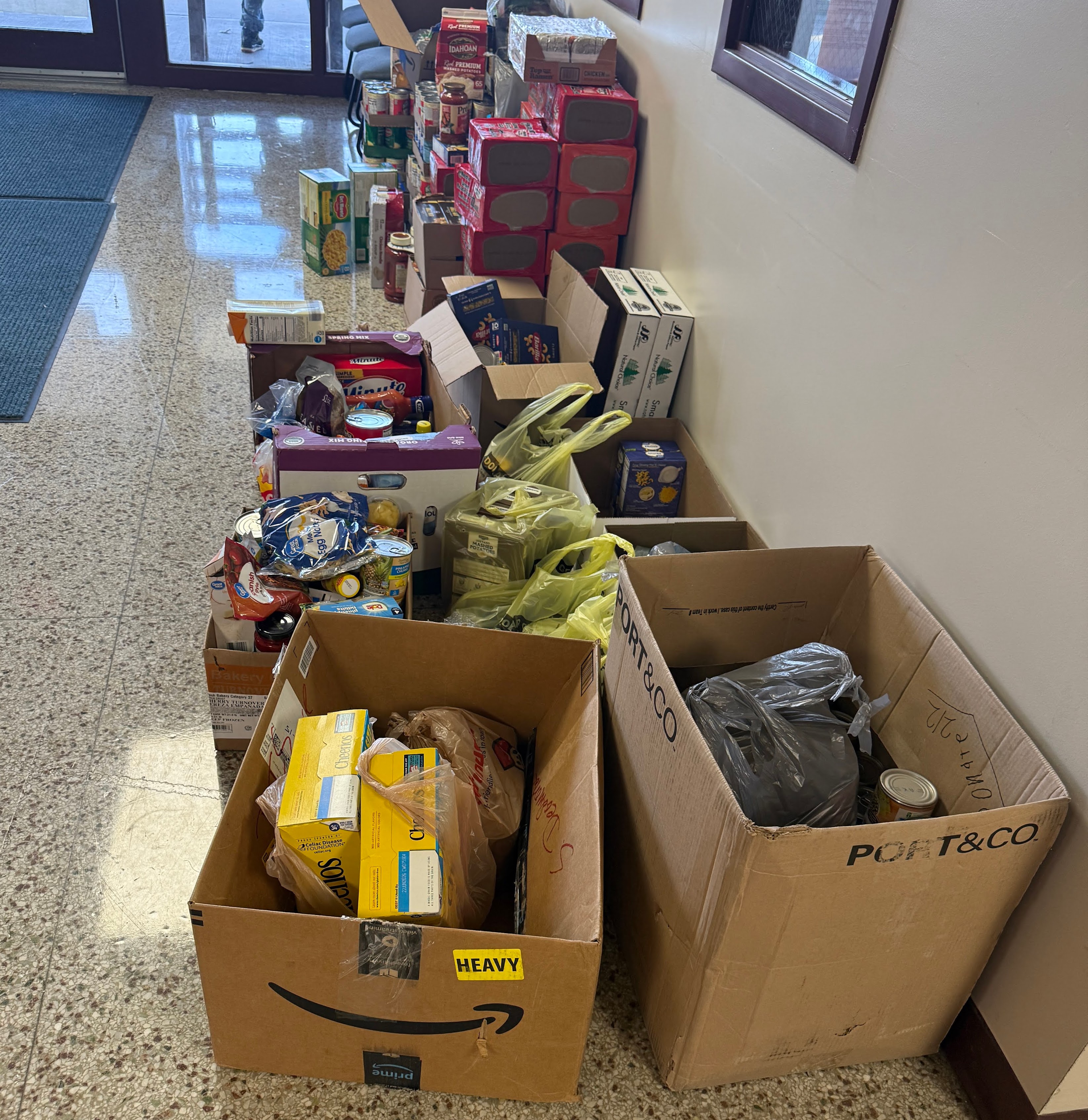 A collection of cardboard boxes and bags filled with non-perishable food items are neatly arranged along a wall.