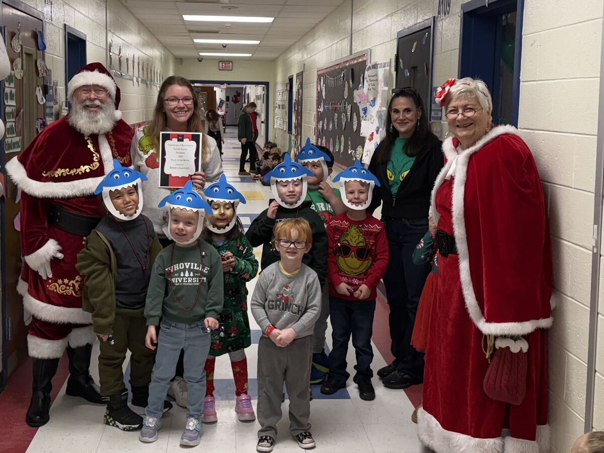 Santa Claus and Mrs. Claus pose with children wearing shark hats in a school hallway decorated for the holidays.