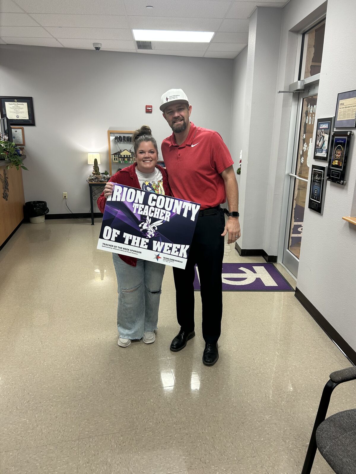 A man and a woman smile while holding a "Marion County Teacher of the Week" sign.