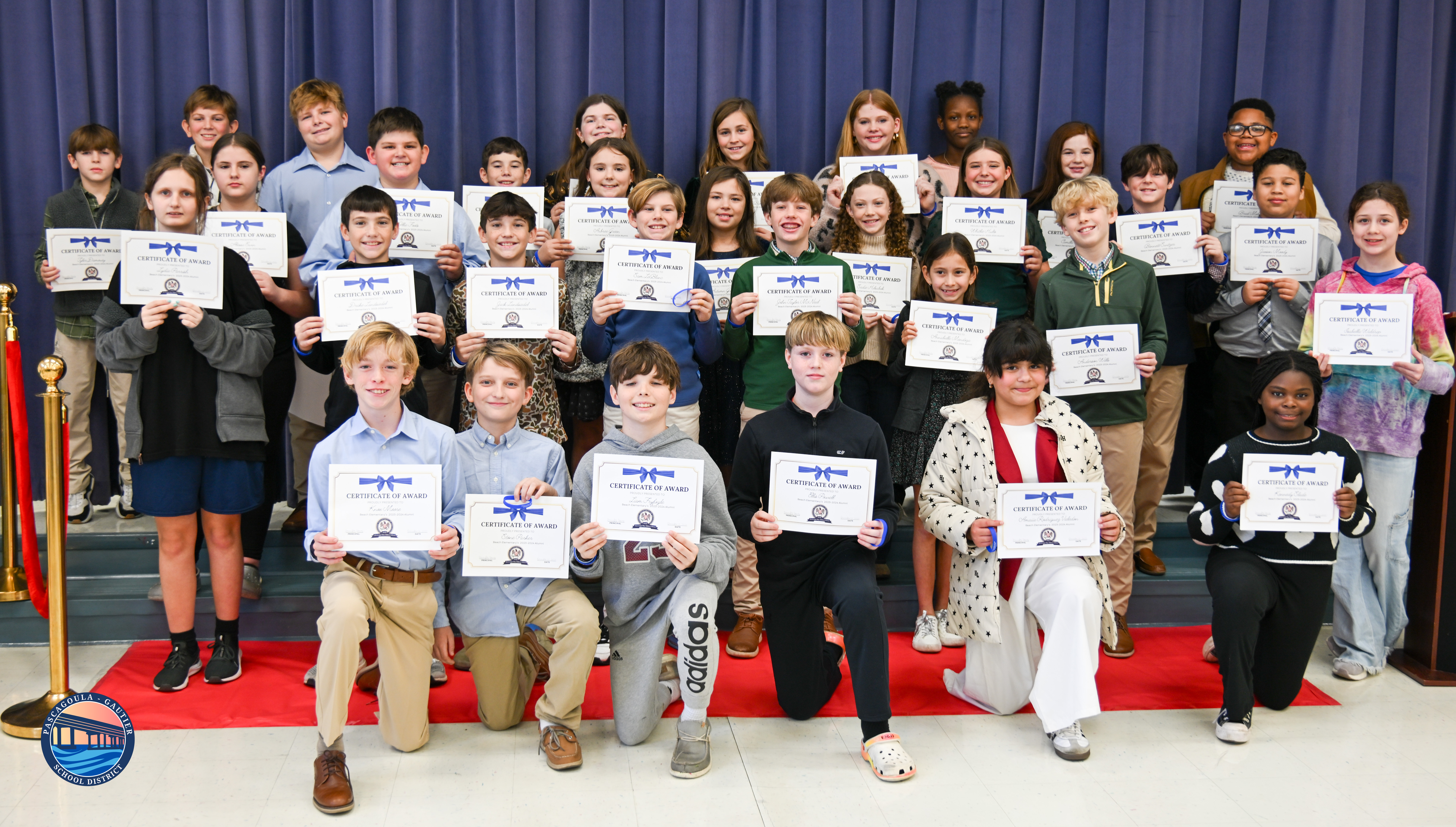 A diverse group of students proudly holds certificates of award on a red carpet in front of a blue curtain.