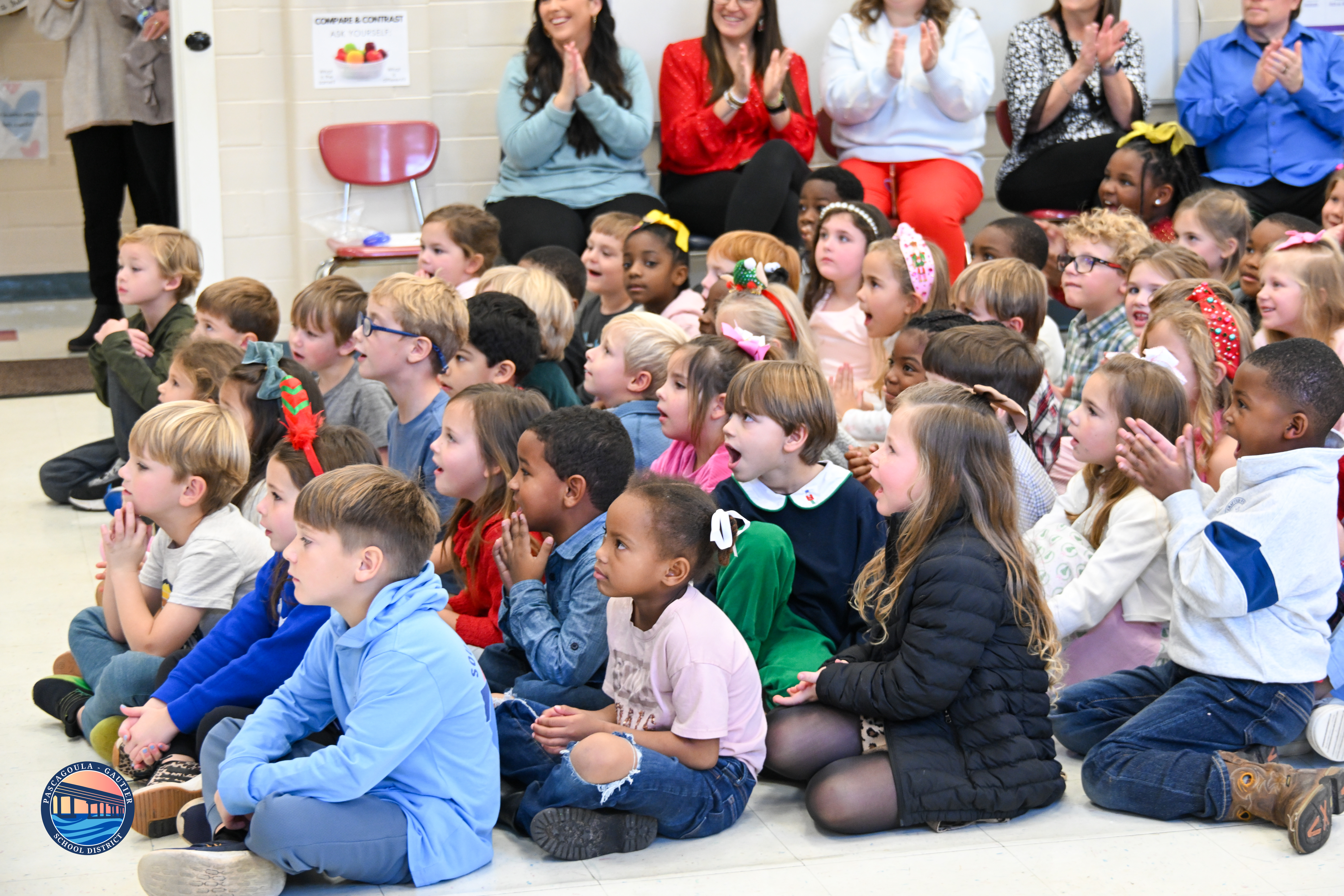 A diverse group of children sit on the floor, attentively watching something in front of them.