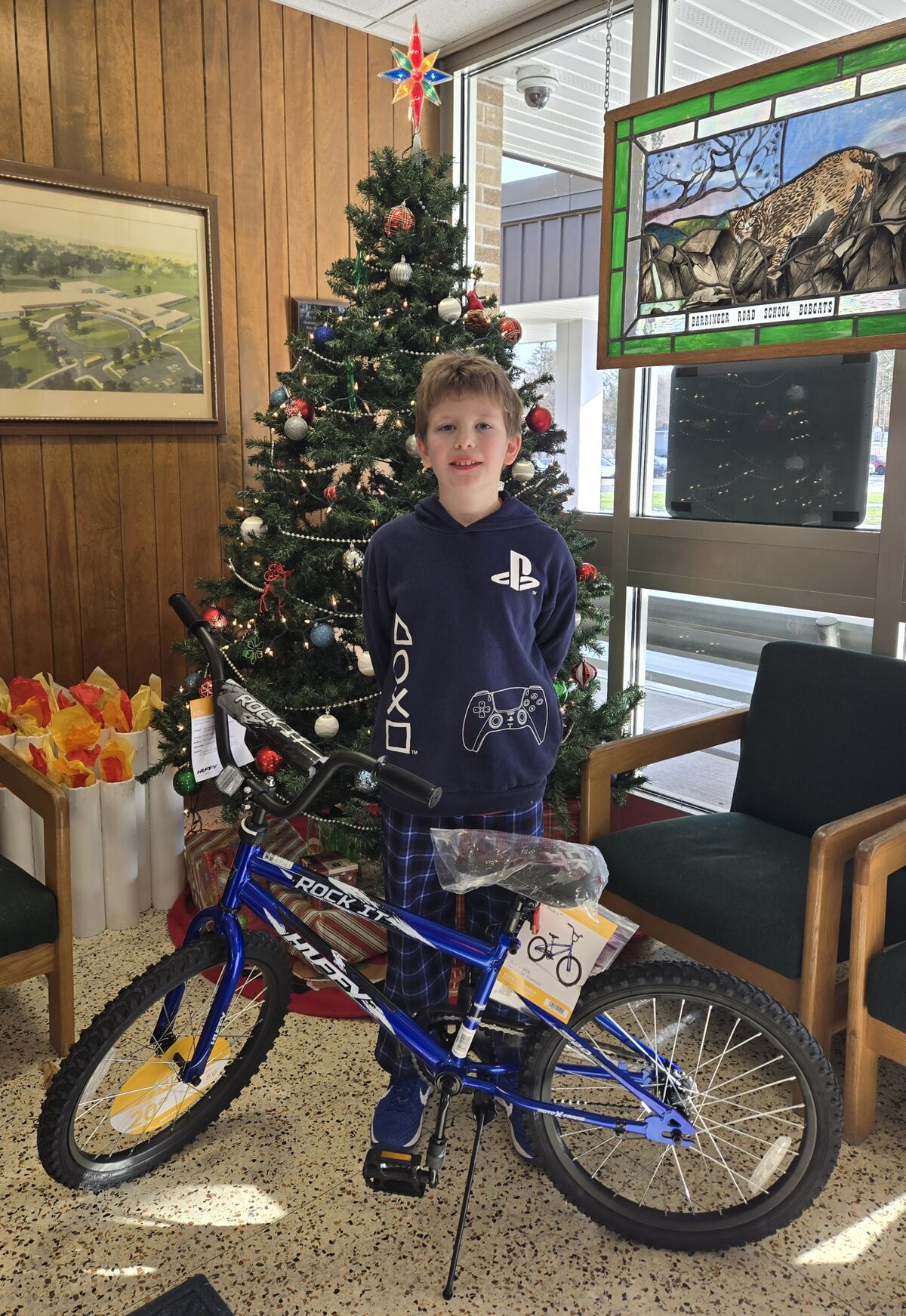 A young boy stands proudly next to a new blue Huffy Rock It bicycle in front of a decorated Christmas tree.