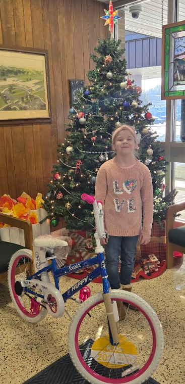 A young girl smiles in front of a decorated Christmas tree, standing next to a new blue Huffy bicycle.