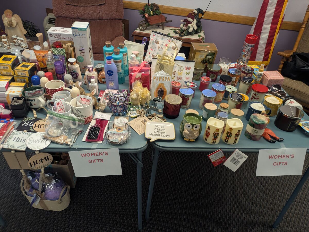 A table displays a variety of items for sale, including lotions, candles, mugs, and decorative owls, with signs indicating 'WOMEN'S GIFTS'.