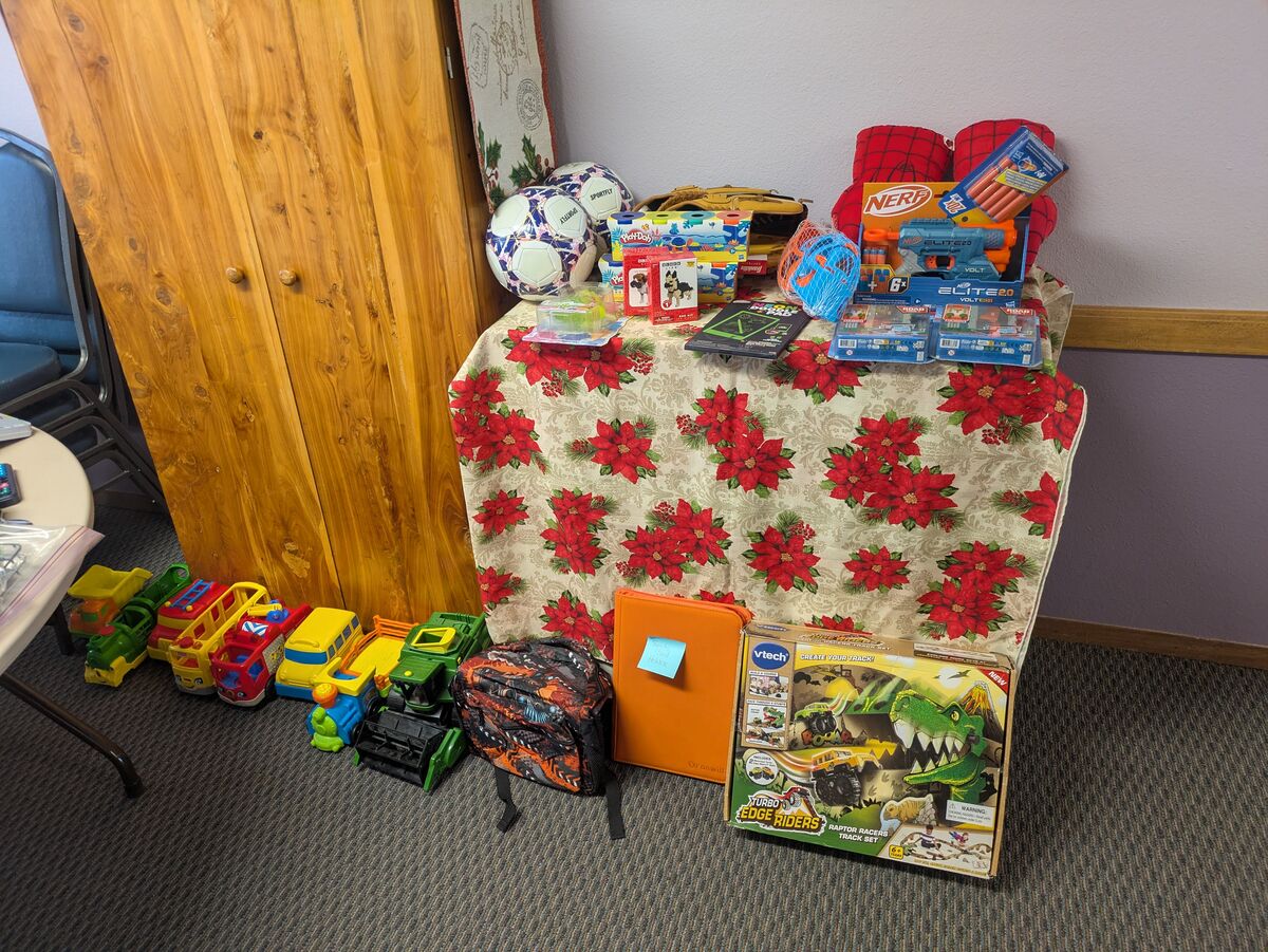 A collection of toys, including toy vehicles, Nerf guns, soccer balls, and a VTech track set, are displayed on a table covered with a poinsettia-patterned cloth.