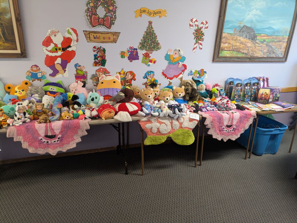 A table is laden with a large collection of stuffed animals and dolls, with holiday decorations on the wall.