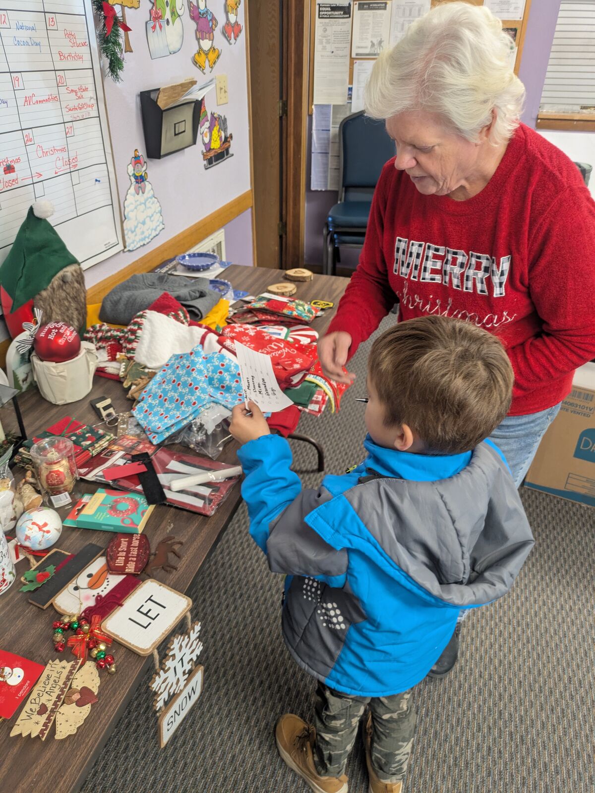 An older woman in a red 'Merry Christmas' sweater looks at a list held by a young boy in a blue jacket.