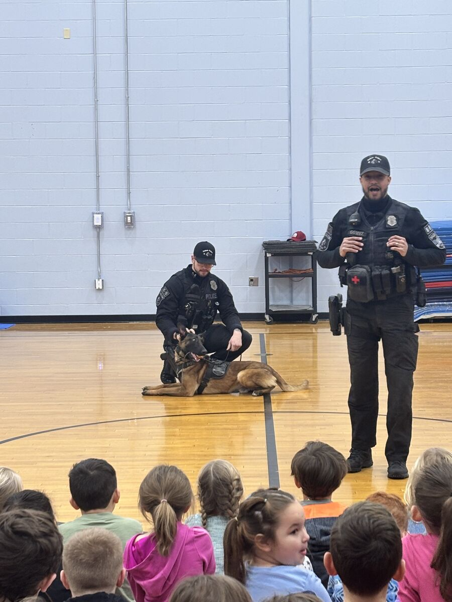 Two police officers and a K9 unit present to a group of children in a gymnasium.