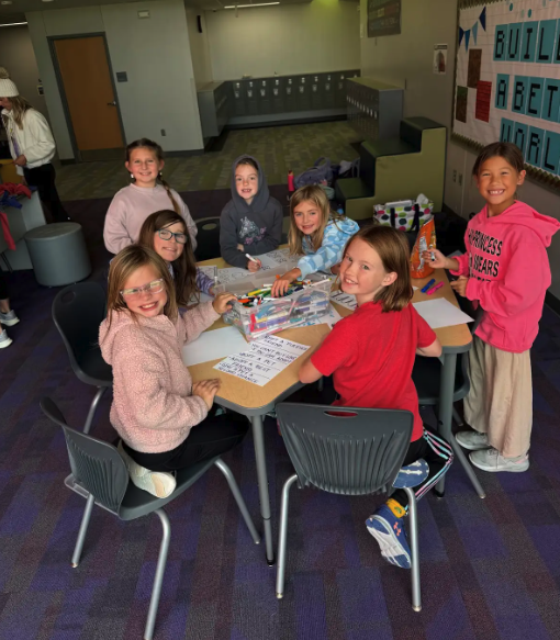 A group of smiling young girls are gathered around a table, engaged in a creative activity with markers and paper.