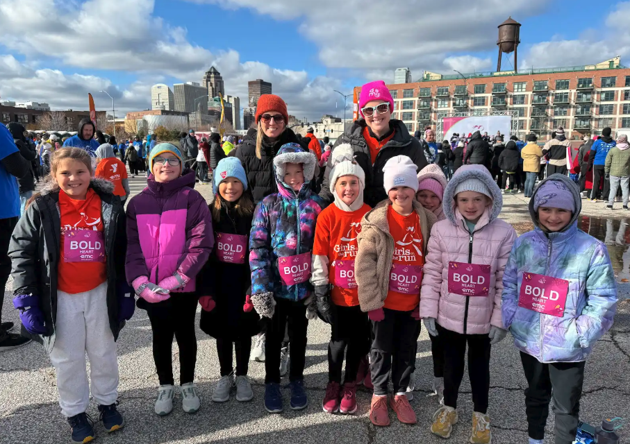 A group of young girls and two adults smile for a photo before a race, wearing winter coats and hats.