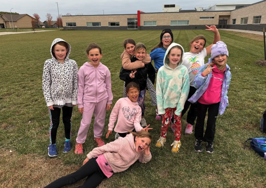 A group of diverse young girls pose for a photo on a grassy field in front of a school building.