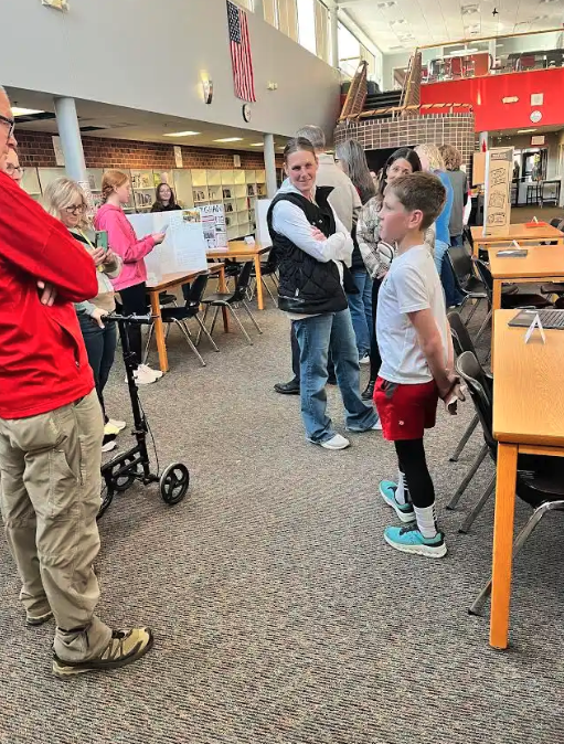 People gather in a school library with displays and tables, an American flag hangs overhead.