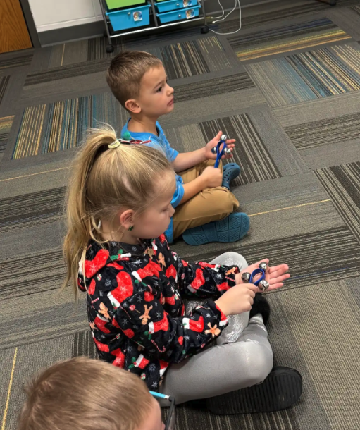 Children sit on a carpeted floor, holding and playing with jingle bells.