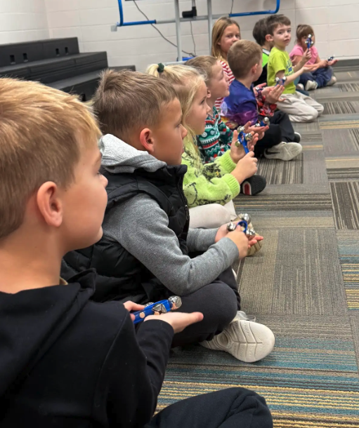 Children sit in a line on the floor, holding musical instruments.
