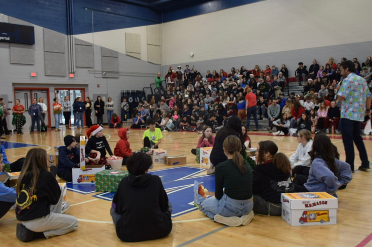 Students sit on a gymnasium floor surrounded by wrapped gifts and boxes, with an audience in the bleachers.