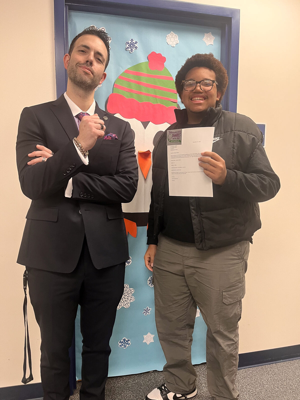A man in a suit and a young person in a puffer jacket stand together, holding a certificate.