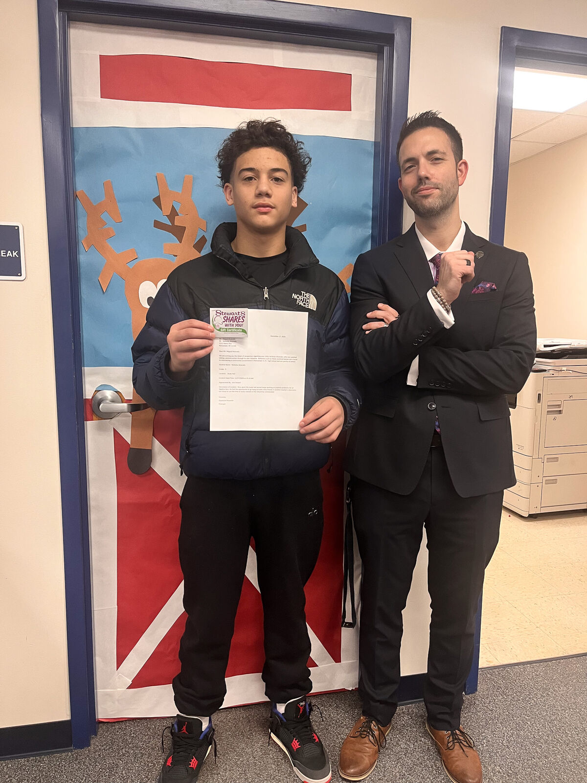 A young person in a puffer jacket holds a gift certificate and a letter, standing next to a man in a suit.