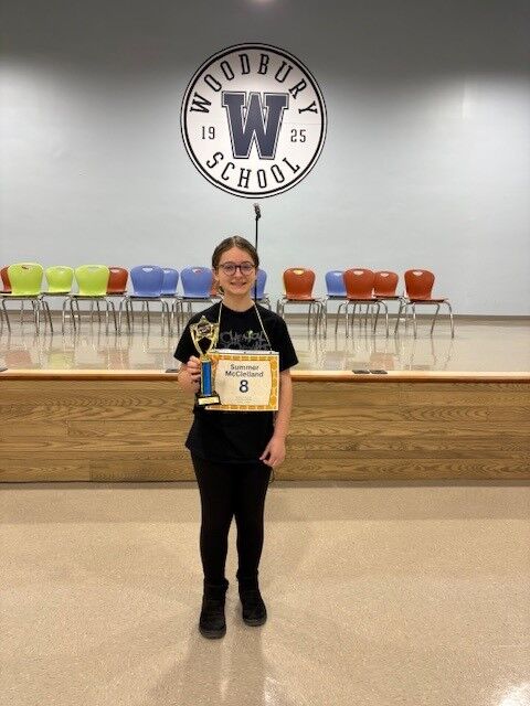 A young person stands on a stage holding a trophy and a certificate, with the Woodbury School logo behind them.