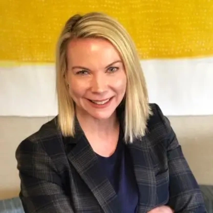 A woman with blonde hair smiles, wearing a plaid blazer over a navy shirt.