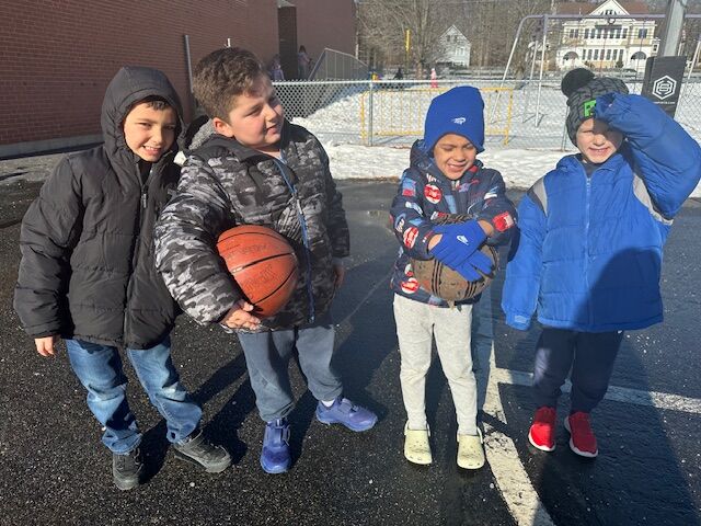 Four young boys in winter jackets stand outdoors with basketballs.