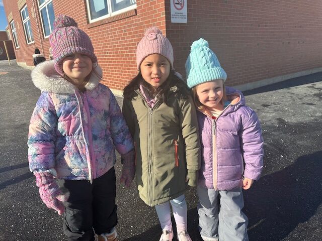 Three young girls in winter coats and hats stand together outdoors.
