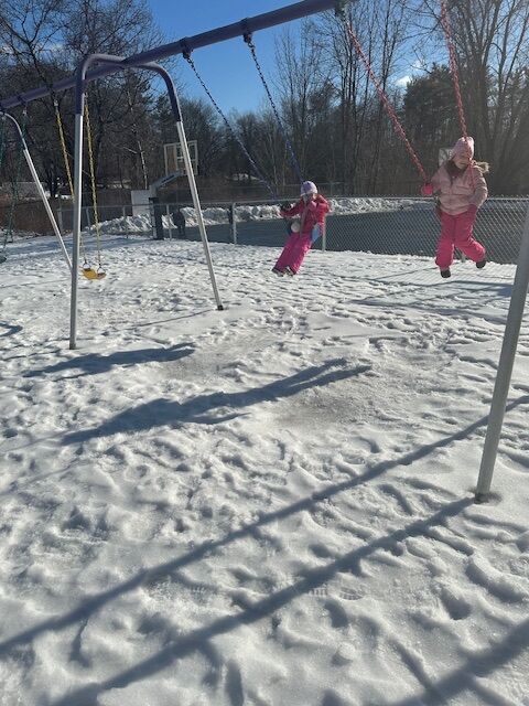 Two children in winter clothing swing on a playground covered in snow.